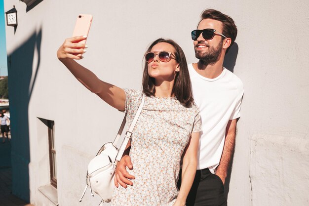 Smiling beautiful woman and her handsome boyfriend Woman in casual summer clothes Happy cheerful family Female having fun Couple posing in the street near wallHugging each other Taking selfie