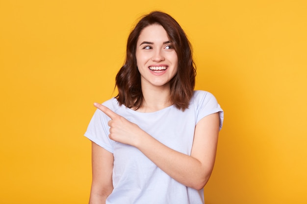 Smiling beautiful young woman wears white casual t shirt, points with index finger aside