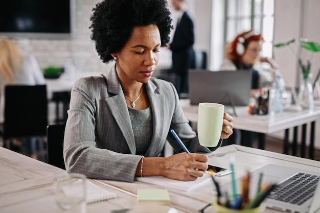 Free photo smiling black businesswoman with cup of coffee working at her desk and taking notes in her notebook there are people in the background