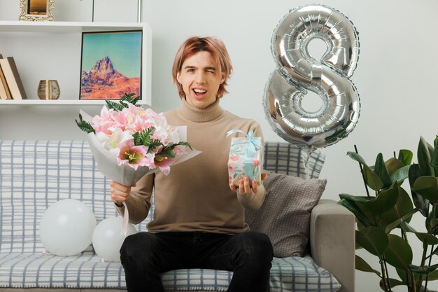 Smiling blinked handsome guy on happy women day holding present with bouquet sitting on sofa in living room