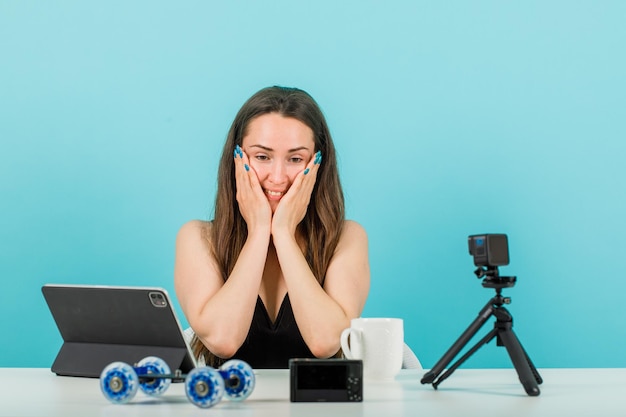 Free photo smiling blogger girl is looking at her little camera by putting hands on cheeks on blue background