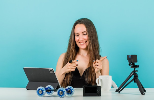 Smiling blogger girl is pointing right with forefingers by looking at tablet screen on blue background