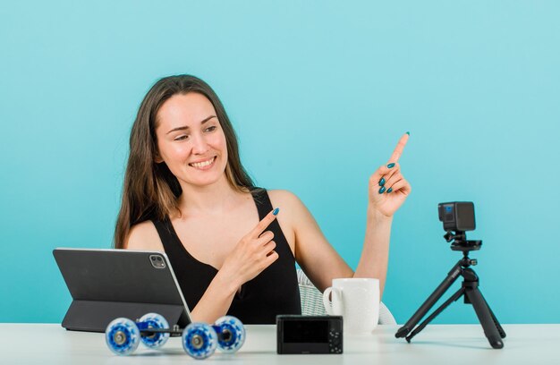 Smiling blogger girl is posing at her little camera by pointing right with forefingers on blue background