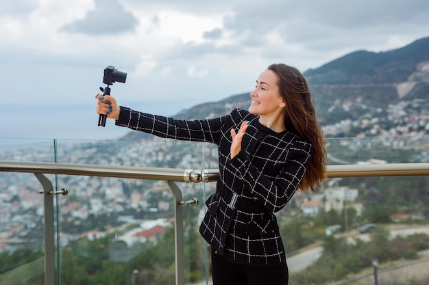 Smiling blogger girl is showing hi gesture to camera in her hand against the background of city view