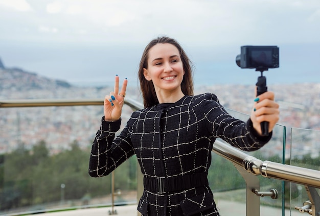 Smiling blogger girl is showing victory gesture to camera in her hand against the background of city view