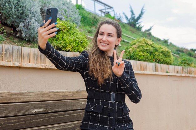 Smiling blogger girl is taking selfie by showing victory gesture by sitting in park