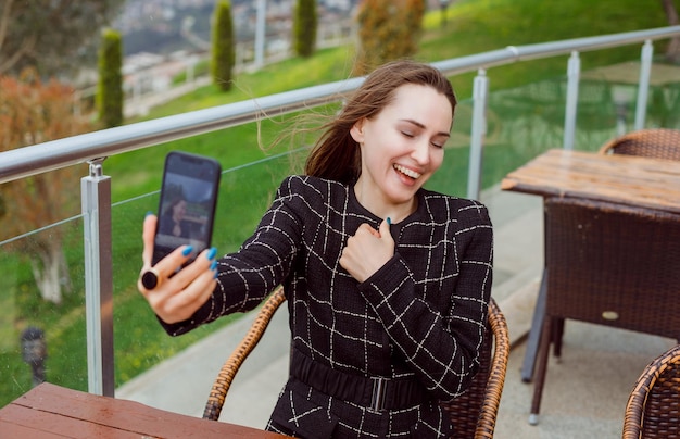 Smiling blogger girl is taking selfie with back camera of smartphone by holding hand on chest on nature view background