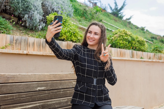 Smiling blogger girl is taking selfie with her smartphone by showing victory gesture in park