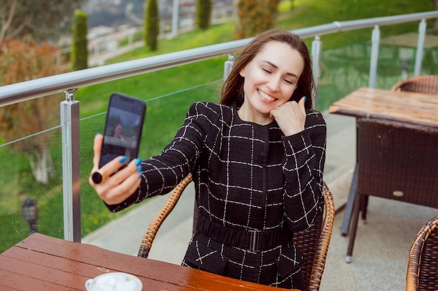 Smiling blogger is taking selfie with back camera of smartphone by holding hand on cheek on nature view background