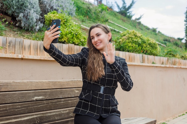 Smiling blogger is taking selfie with her smartphone by showing hi gesture in park