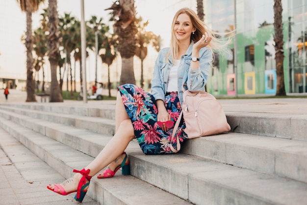 Free photo smiling blond woman sitting on stairs in city street in stylish printed skirt and denim oversize jacket with leather backpac