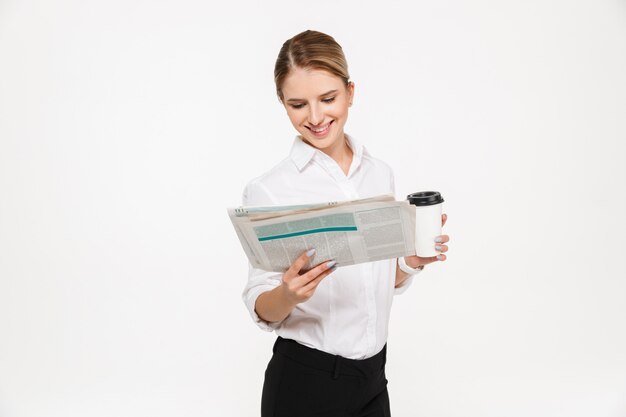 Smiling blonde business woman reading newspaper while holding cup of coffee over white wall