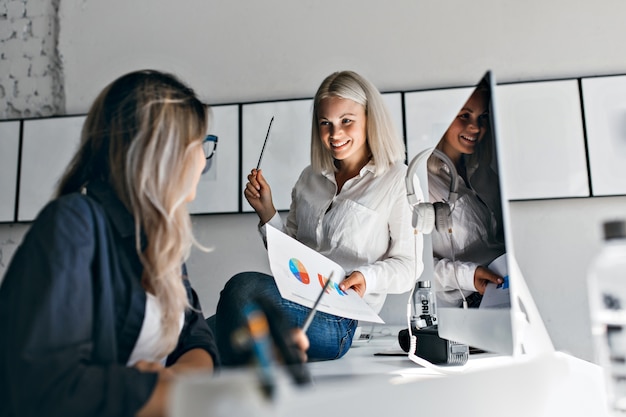 Free Photo smiling blonde female manager holding infographic and pencil, while sitting on table. indoor portrait of two women working with computer in office.