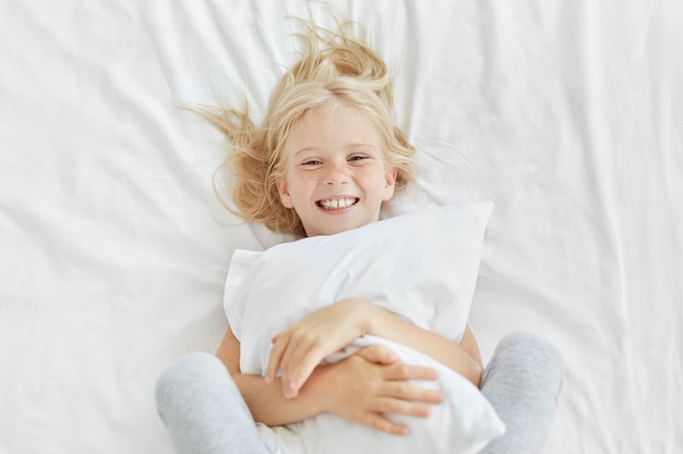 Free photo smiling blonde girl embracing white pillow while being in kindergarten, having good mood while seeing someone and lying in white bed. little adorable female child having beddtime. rest concept