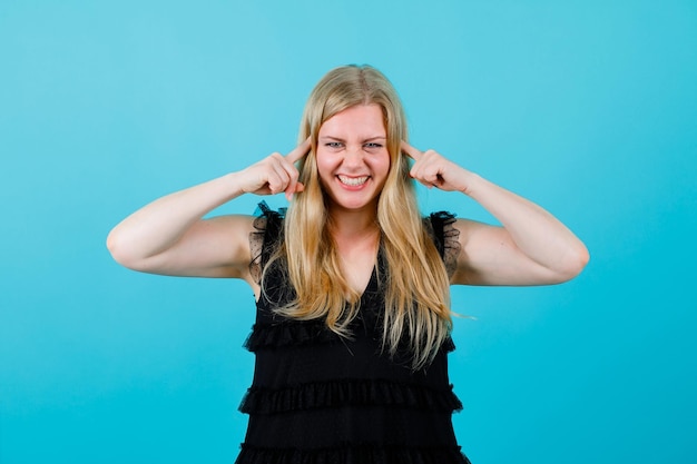 Smiling blonde girl is covering her ears with forefingers on blue background