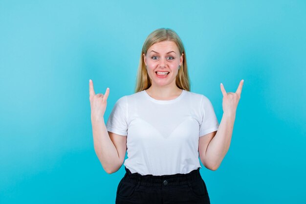 Smiling blonde girl is hsowing rock gestures on blue background