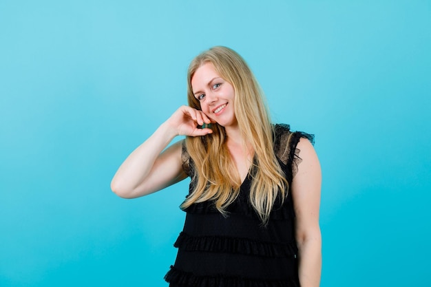 Smiling blonde girl is looking away by holding hand cheek on blue background