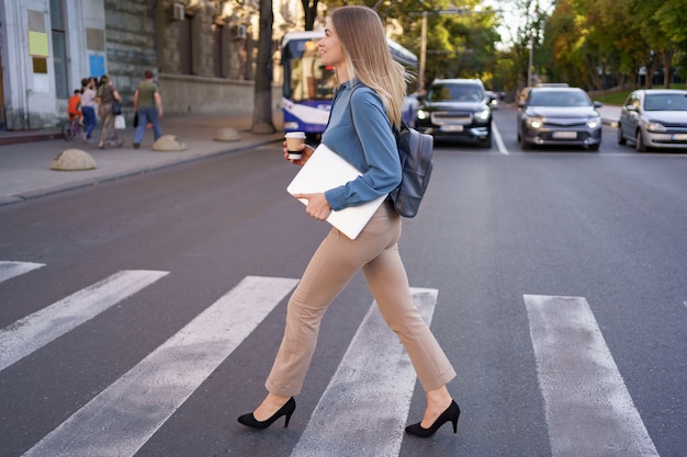 Smiling blonde woman crossing zebra with coffee and laptop