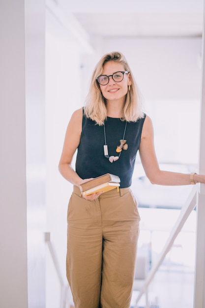 Smiling blonde young woman holding books in hand looking at camera