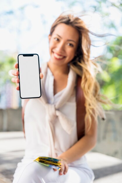 Smiling blonde young woman holding map in hand showing mobile blank white screen