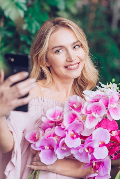 Smiling blonde young woman holding orchid in hand taking self portrait from mobile phone