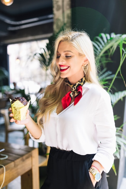 Smiling blonde young woman with hand on pocket holding muffin in paper holder