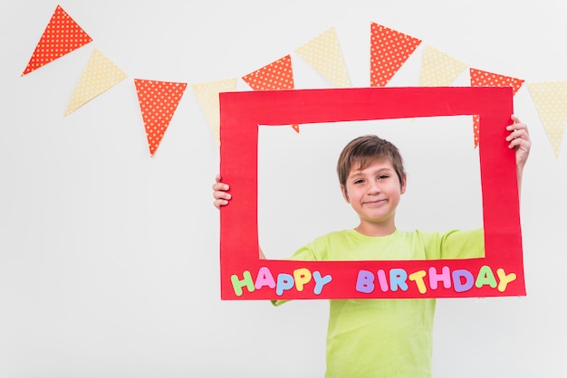 Free photo smiling boy holding frame with happy birthday frame against wall decorated with bunting