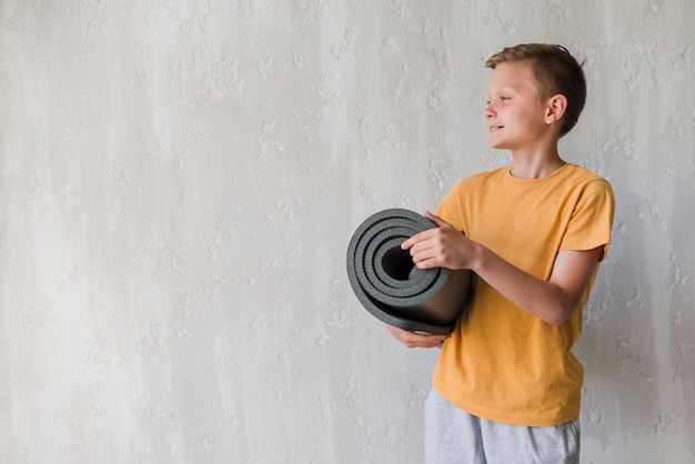 Free Photo smiling boy holding rolled up exercise mat looking away in front of concrete wall