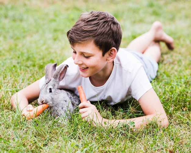 Free photo smiling boy lying on green grass feeding carrot to rabbit