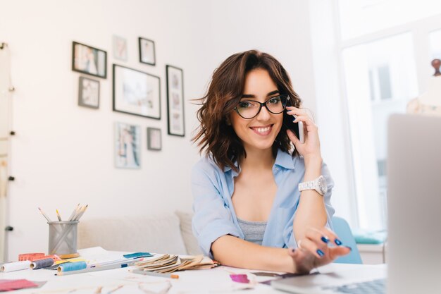 A smiling brunette girl in a blue shirt is working in the workshop.  She is speaking on phone and typing on computer. She is smiling to the camera.