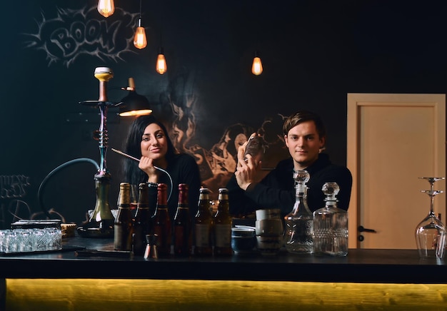 Smiling brunette girl with hookah and barmen at the counter in a nightclub, looking at camera.