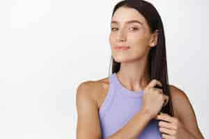 Free photo smiling brunette woman brushing her healthy long dark hair adjusting hairstyle standing in tanktop against white background