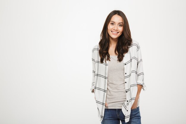 Smiling brunette woman in shirt posing with arms in pockets and looking at the camera over gray