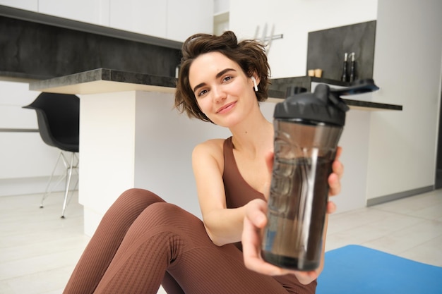 Free Photo smiling brunette woman sits on yoga fitness mat gives you water bottle to drink after workout does p