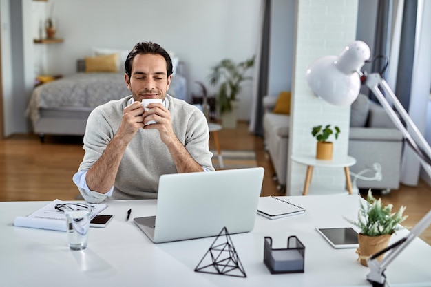 Smiling businessman enjoying in coffee break with his eyes closed while working at home