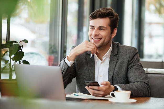 Smiling businessman sitting by the table in cafe with laptop computer and smartphone while looking away