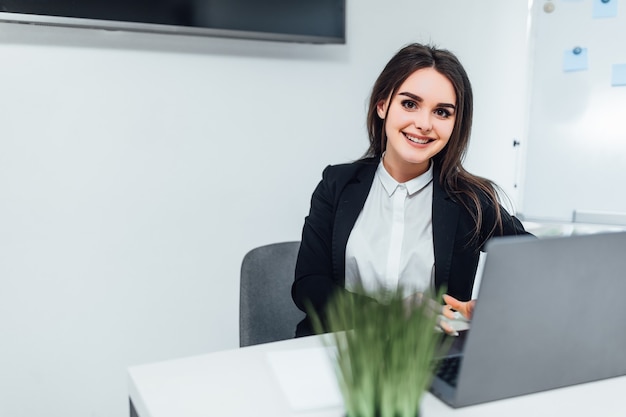 Smiling businesswoman dressed casual suit sitting in modern office and using laptop. With copy space.