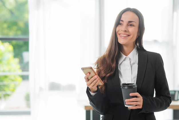 Smiling businesswoman holding smartphone and disposable coffee cup