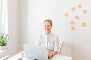 Free photo smiling businesswoman using laptop with sticky notes on wall