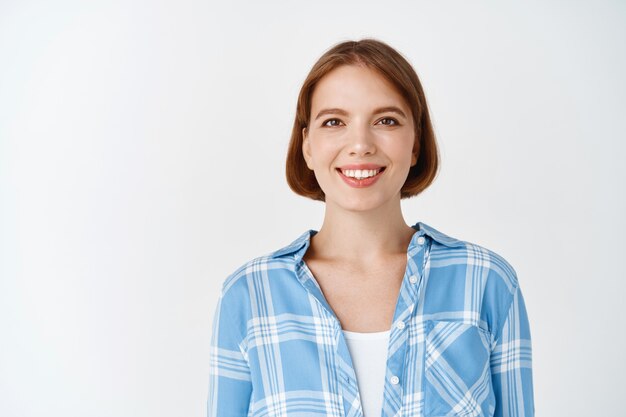 Smiling candid woman in casual clothes looking happy. Young girl with short hair and light make-up standing on white wall