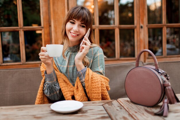 Smiling carefree  woman have a coffee break in cozy cafe with wood interior , using mobile phone. Holding cup of hot cappuccino. Winter season.