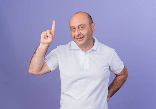 Smiling casual mature businessman keeping hand behind back and raising finger isolated on purple background