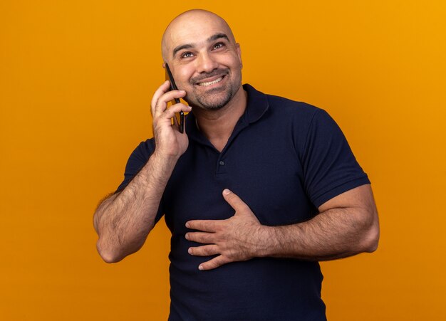 Smiling casual middle-aged man keeping hand on belly looking up talking on phone isolated on orange wall