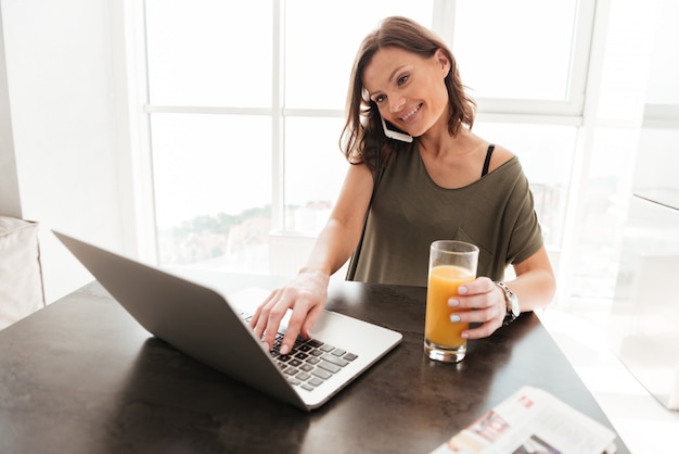 Smiling casual woman talking on smartphone, using laptop computer and drinking juice by the table