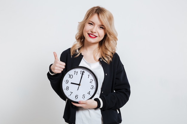 Free photo smiling casual young woman teenager holding clock and showing thumb up