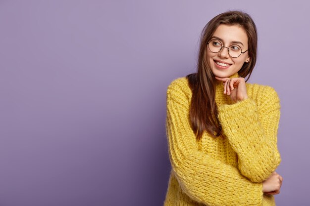 Smiling Caucasian female model touches gently chin, looks with toothy smile aside