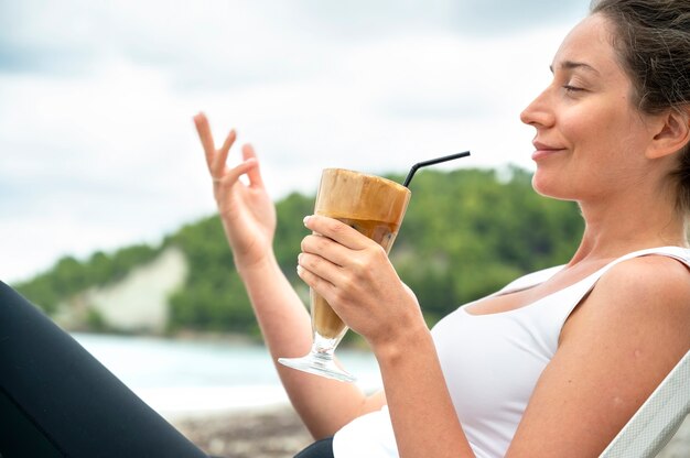 Smiling caucasian woman holding coffee drink on a beach with foam and drinking straw with hills