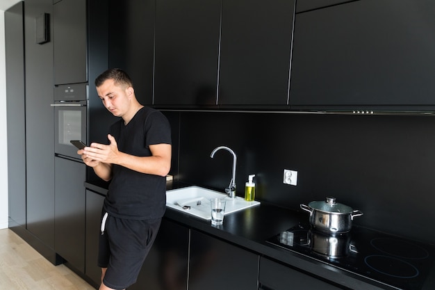 Free photo smiling caucasian young man use cellphone gadget texting messaging standing in kitchen