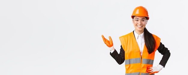 Smiling cheerful asian female construction manager introduce her interprise for investors wearing safety glasses helmet and gloves pointing hand left and looking proud white background