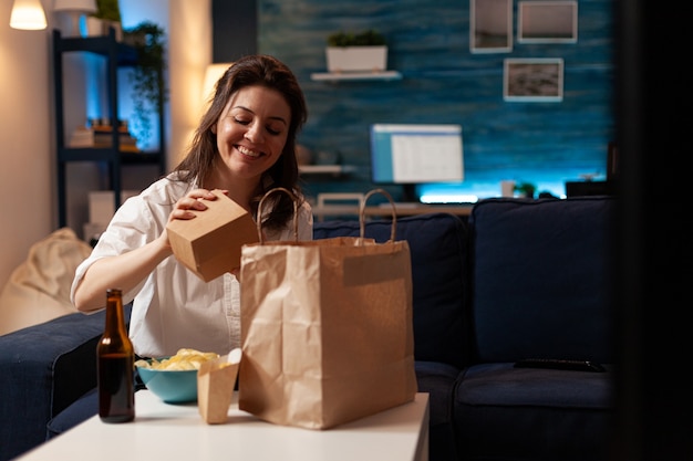 Free Photo smiling cheerful woman unpacking tasting fastfood home delivered sitting on couch